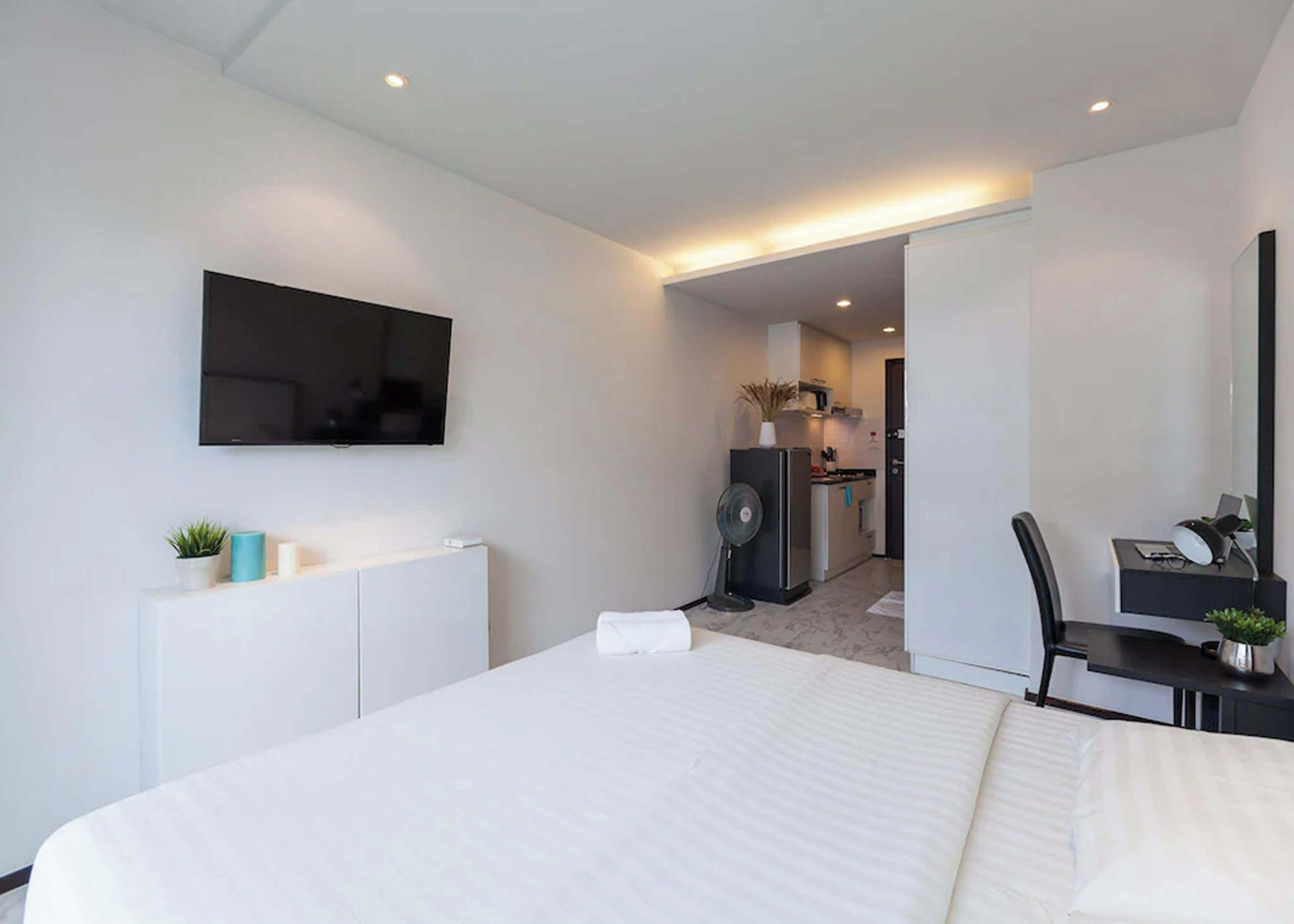 A modern hotel room with a large bed, a flat-screen TV mounted on the wall, a desk with a chair, and a kitchenette in the background.