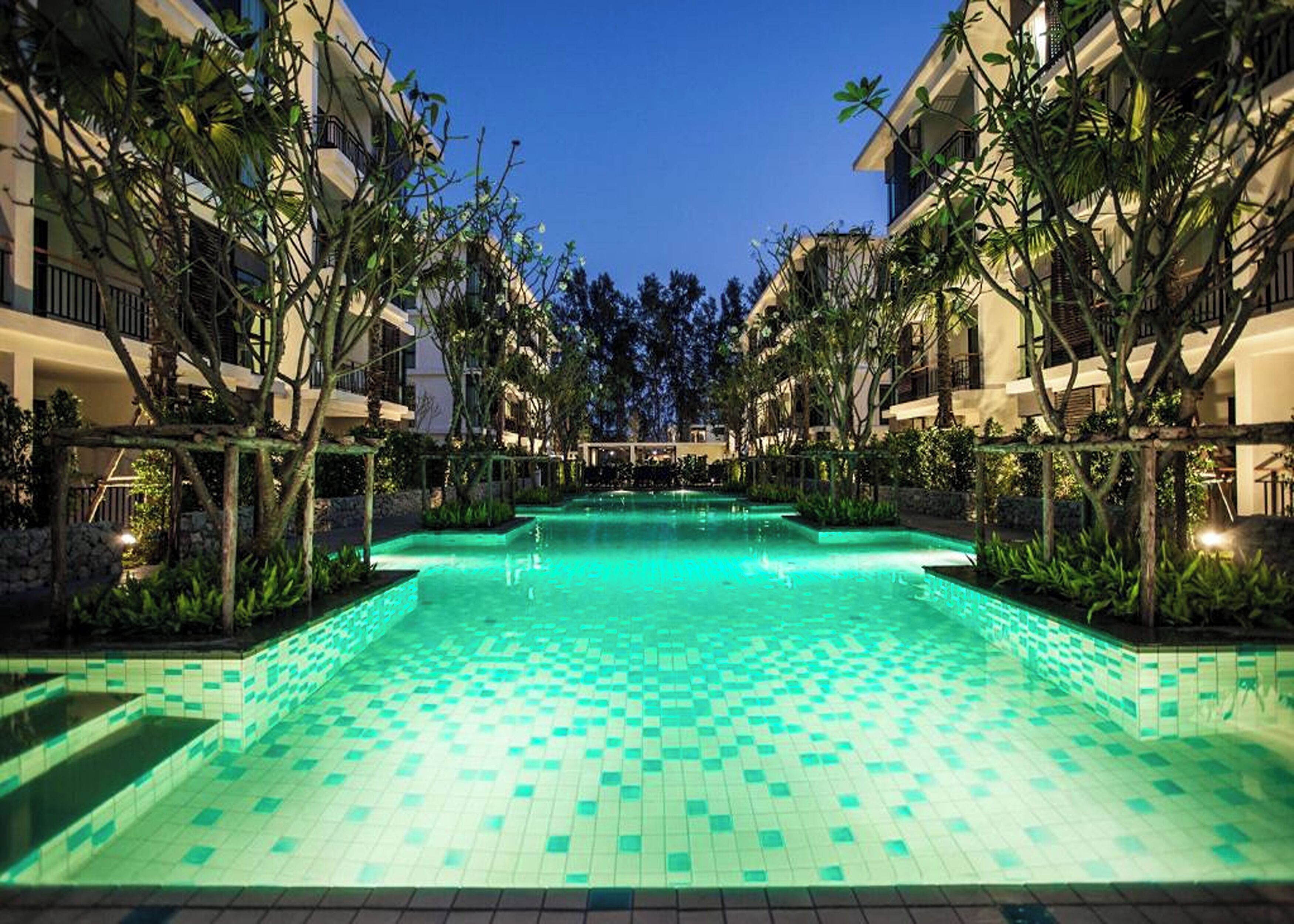 Una piscina con suelo de baldosas, rodeada de abundante vegetación y edificios de varios pisos con balcones.