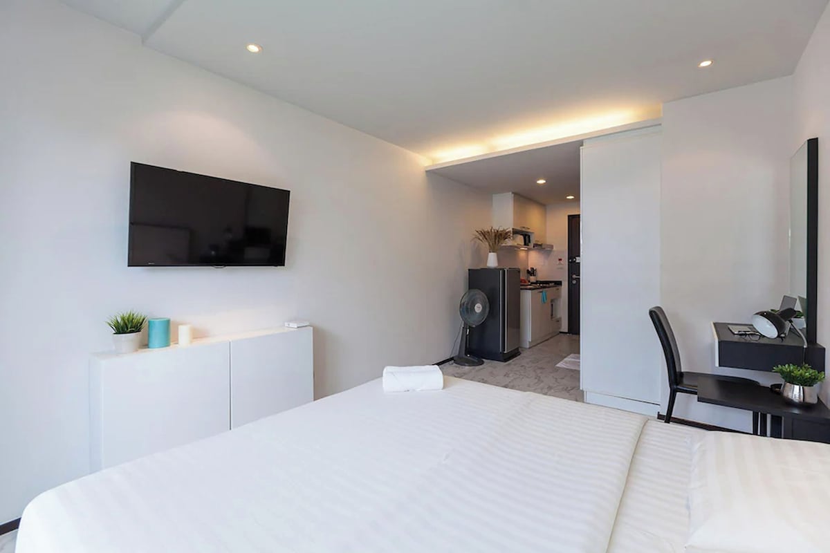 Una habitación de hotel moderna con una cama grande, un televisor de pantalla plana, un escritorio con silla y una pequeña kitchenette al fondo.