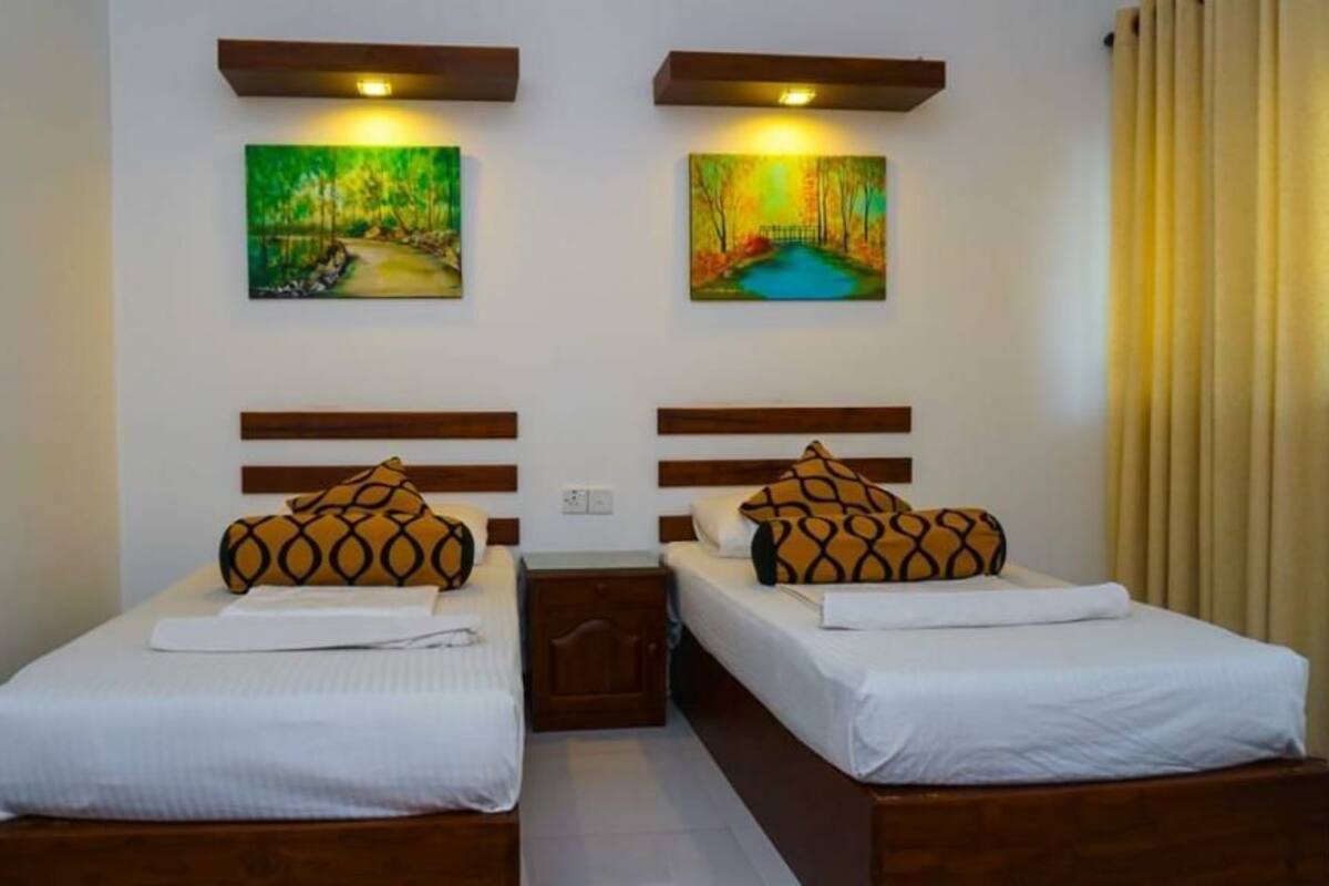 Een hotelkamer met twee bedden, elk met een houten hoofdbord en een witte spreidek. Boven de bedden hangen twee schilderijen, en daartussen staat een nachtkastje.