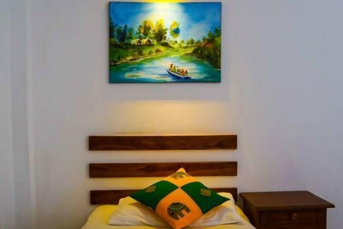 Een netjes opgemaakt bed met een groen en oranje kussen, erboven een schilderij van een rivierlandschap, en een houten hoofdbord.