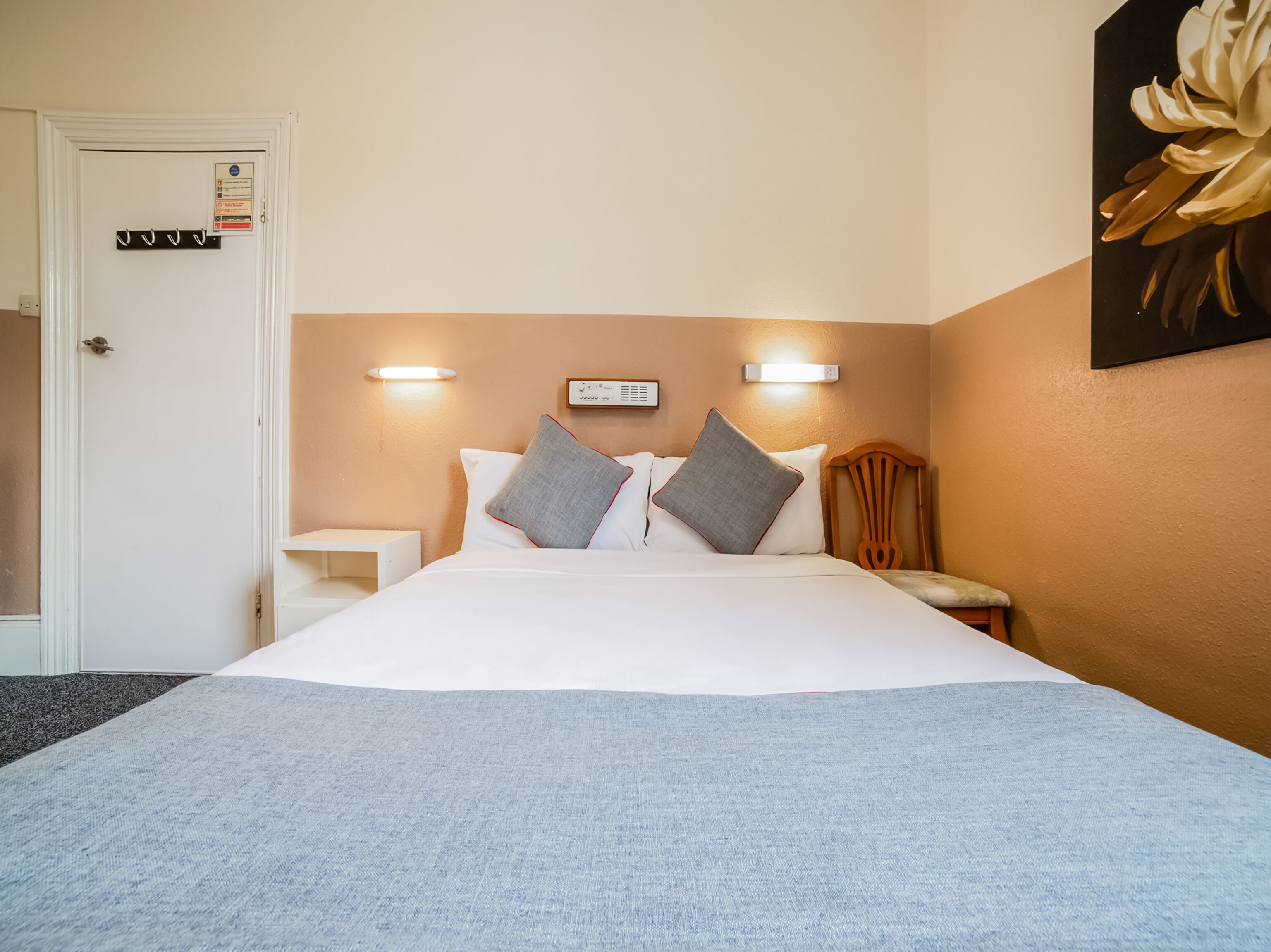 A hotel room with a bed, two bedside tables, a nightstand, a wall-mounted light, and a door with a sign.