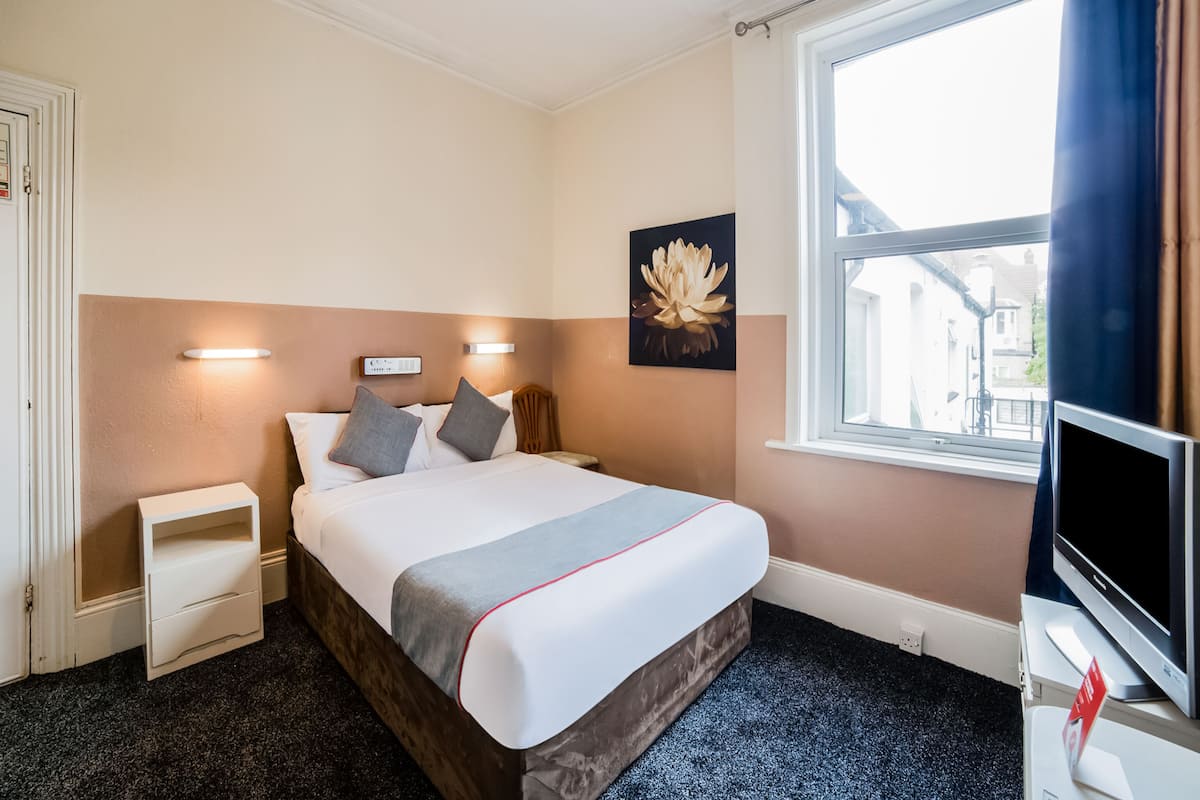 Une chambre d’hôtel équipée d’un lit, d’une table de chevet, d’une télévision et d’une fenêtre donnant sur des immeubles.