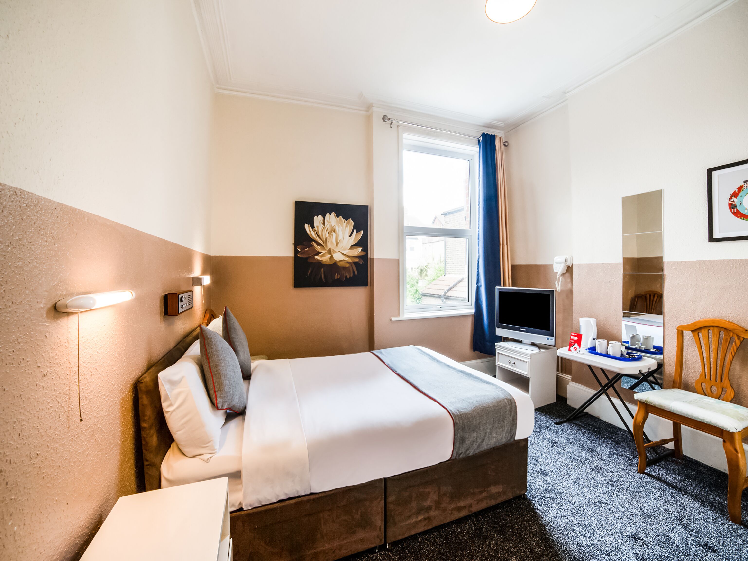 A hotel room with a bed, a desk with a computer, a chair, a window with curtains, and a wall-mounted artwork.