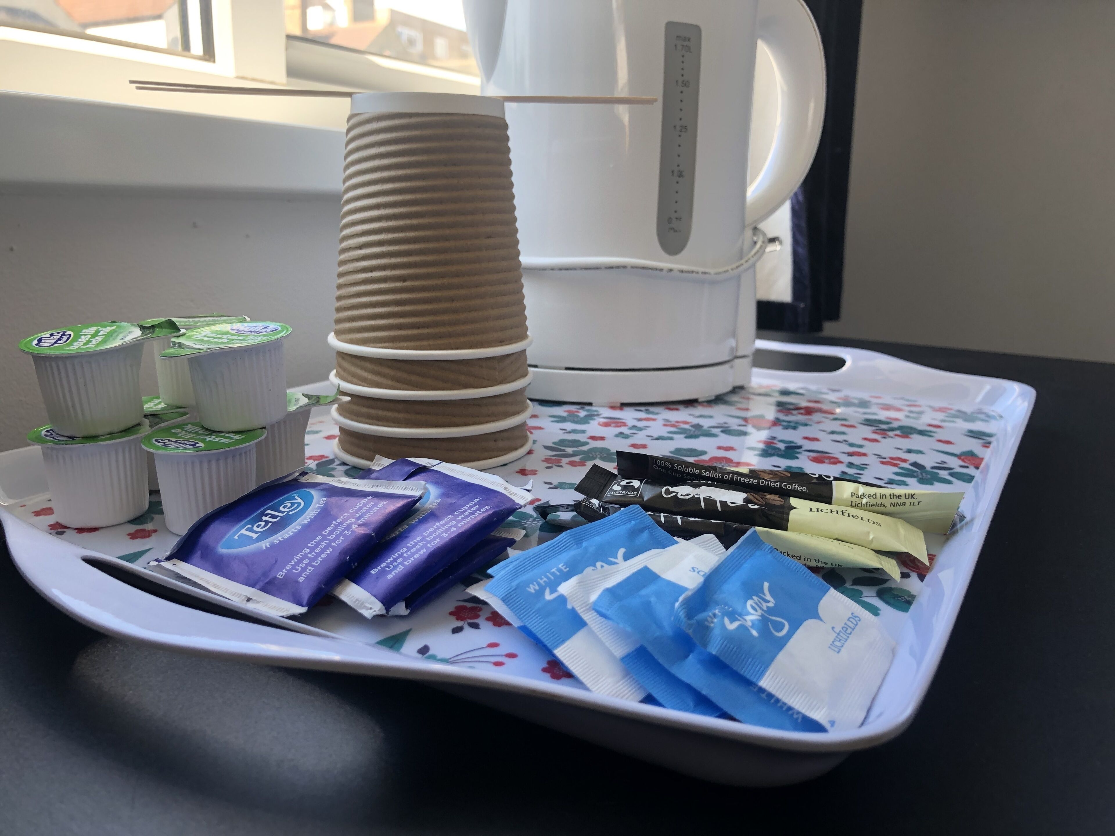 A tray with coffee pods, a stack of paper cups, and a white coffee maker on a floral tablecloth.