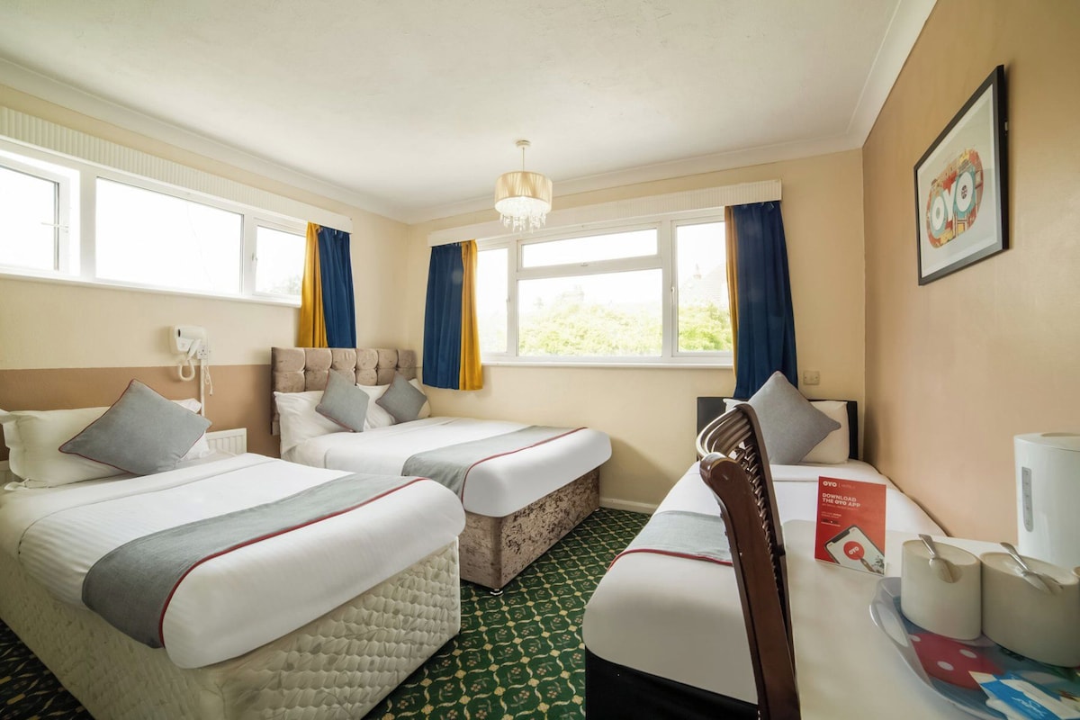A hotel room with two beds, a desk, a chair, a window with curtains, a chandelier, and a framed picture on the wall.