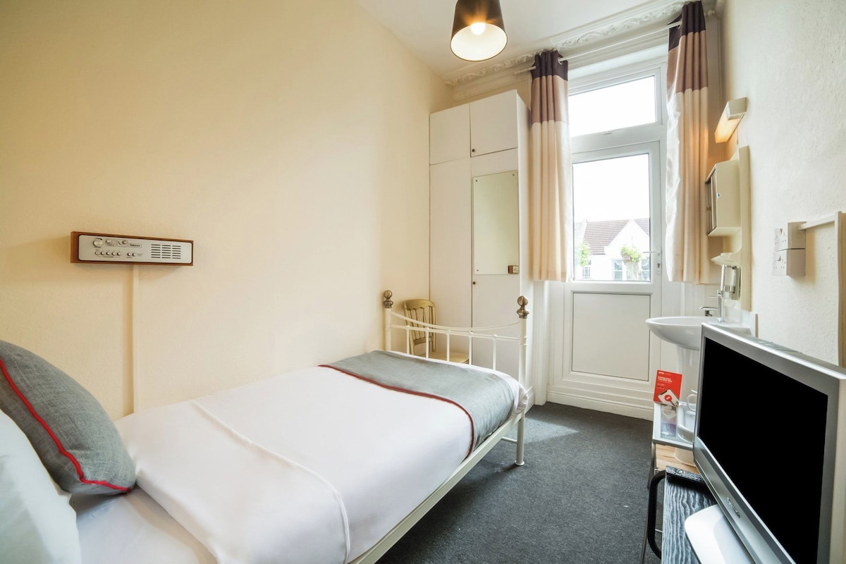 A single bed with a headboard, a bedside table, a television, and a window with curtains.