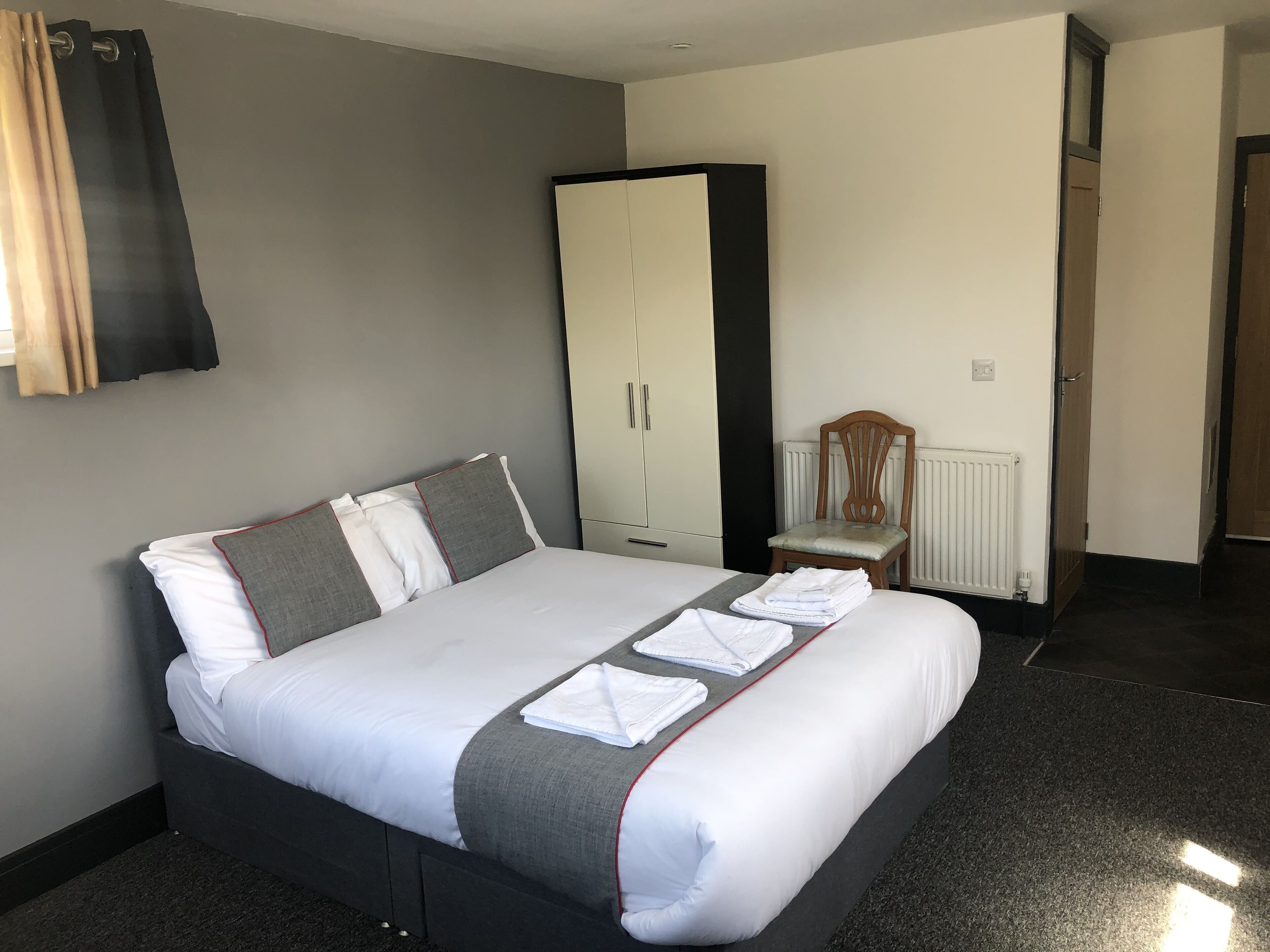 A neatly made bed with white and grey bedding, a wooden chair, a wardrobe, and a radiator.