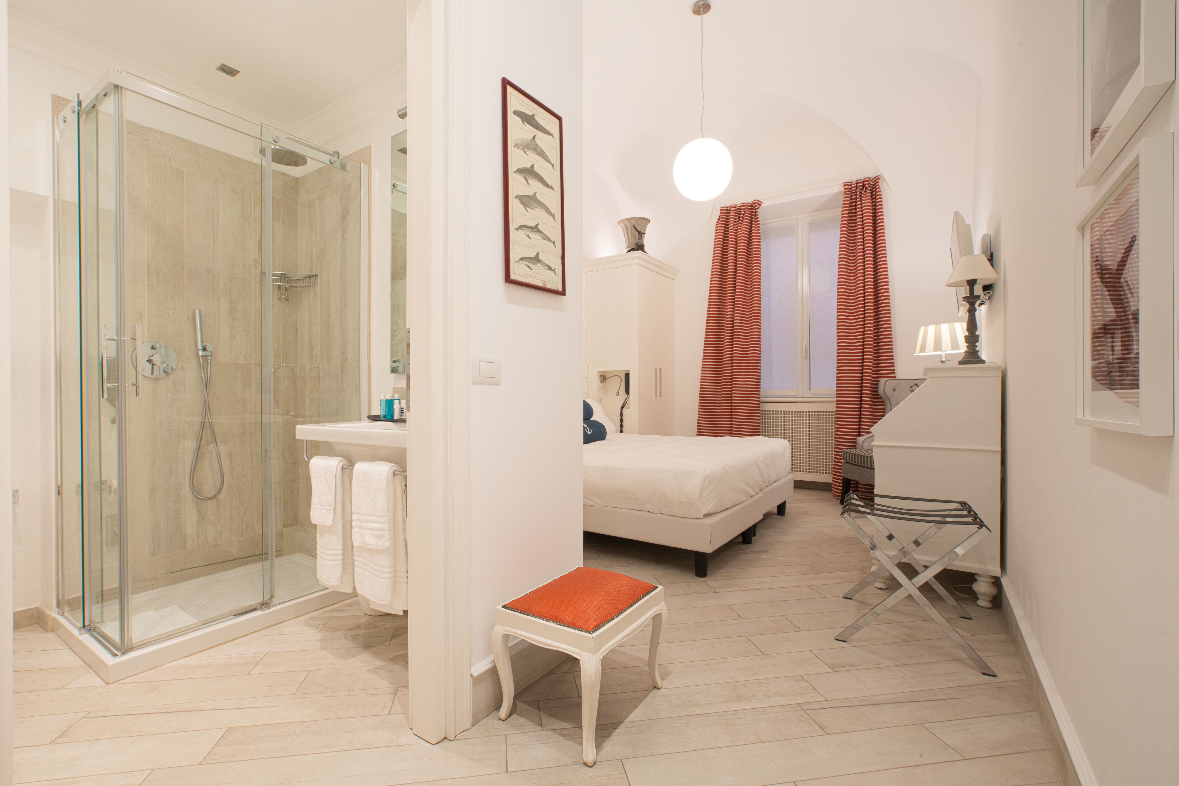 Ein modernes Badezimmer mit einer gläsernen Duschkabine, einem weißen Waschbecken, einem kleinen orangefarbenen Hocker und einem Bett mit weißer Matratze.