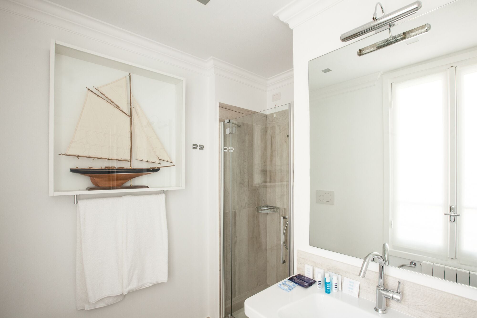 Un bagno con un grande specchio, un modellino di barca a vela in legno, un box doccia in vetro e articoli da toilette sul lavandino.