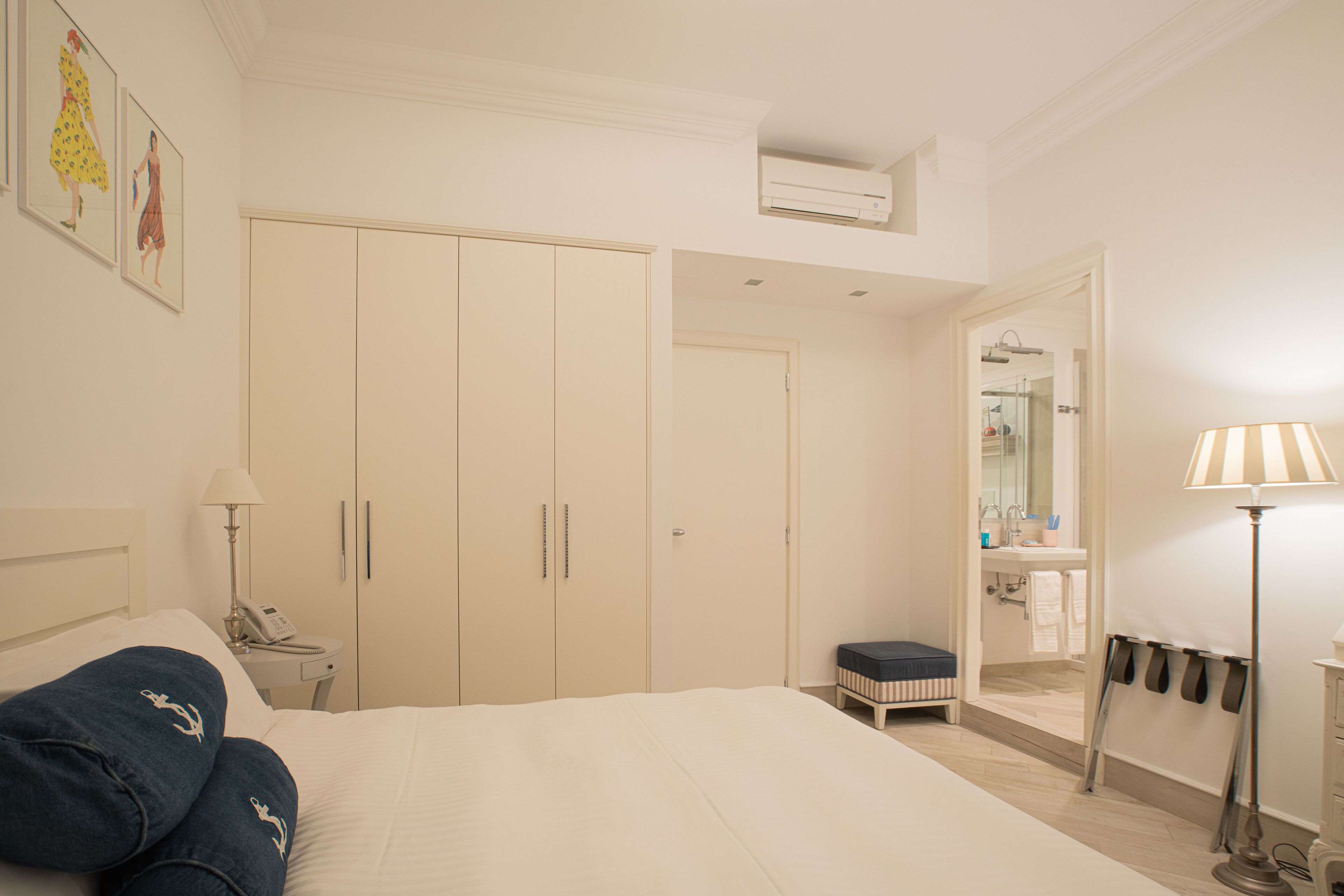 A hotel room with a bed, a bedside table with a lamp, a wardrobe, a bathroom visible through an open door, and a small stool.
