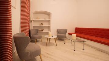 A modern living room with a red sofa, two grey armchairs, a glass coffee table, and a white shelving unit against a white wall.