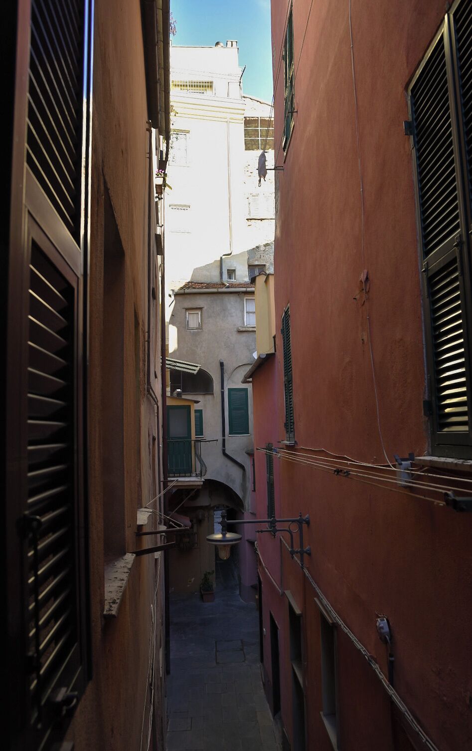 Un estrecho callejón entre edificios con contraventanas, un pequeño puente y una torre con campanas.