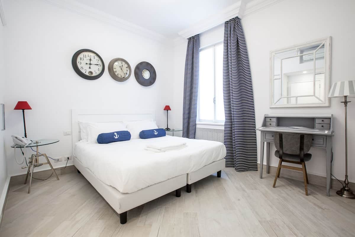 A hotel room with a bed, a desk, a chair, a lamp, a telephone, and three clocks on the wall.
