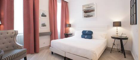 A hotel room with a large bed, a chair, a bedside table with a lamp, and a wall-mounted shelf with decorative items.