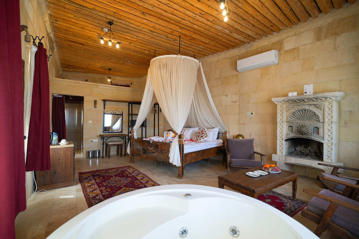 A bedroom with a large bed, a wooden ceiling, a fireplace, and a bathroom with a bathtub.