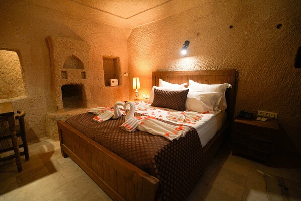 A room with a stone wall, a wooden bed with a headboard, a nightstand, a lamp, and a small shelf. - Helike Cave Suites (Nevsehir)
