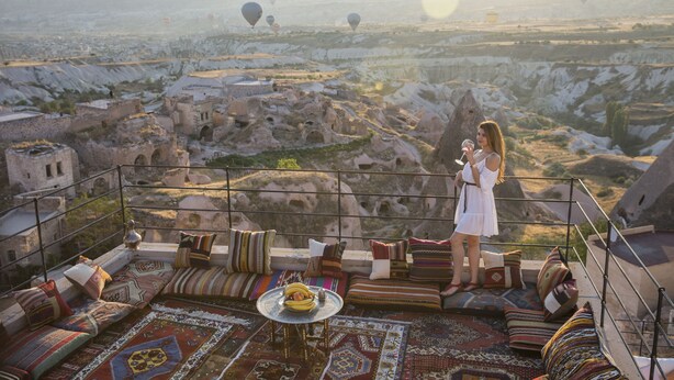 A terrace with a view of a mountainous landscape, furnished with colorful cushions and a small table with a fruit bowl. - Helike Cave Suites (Nevsehir)