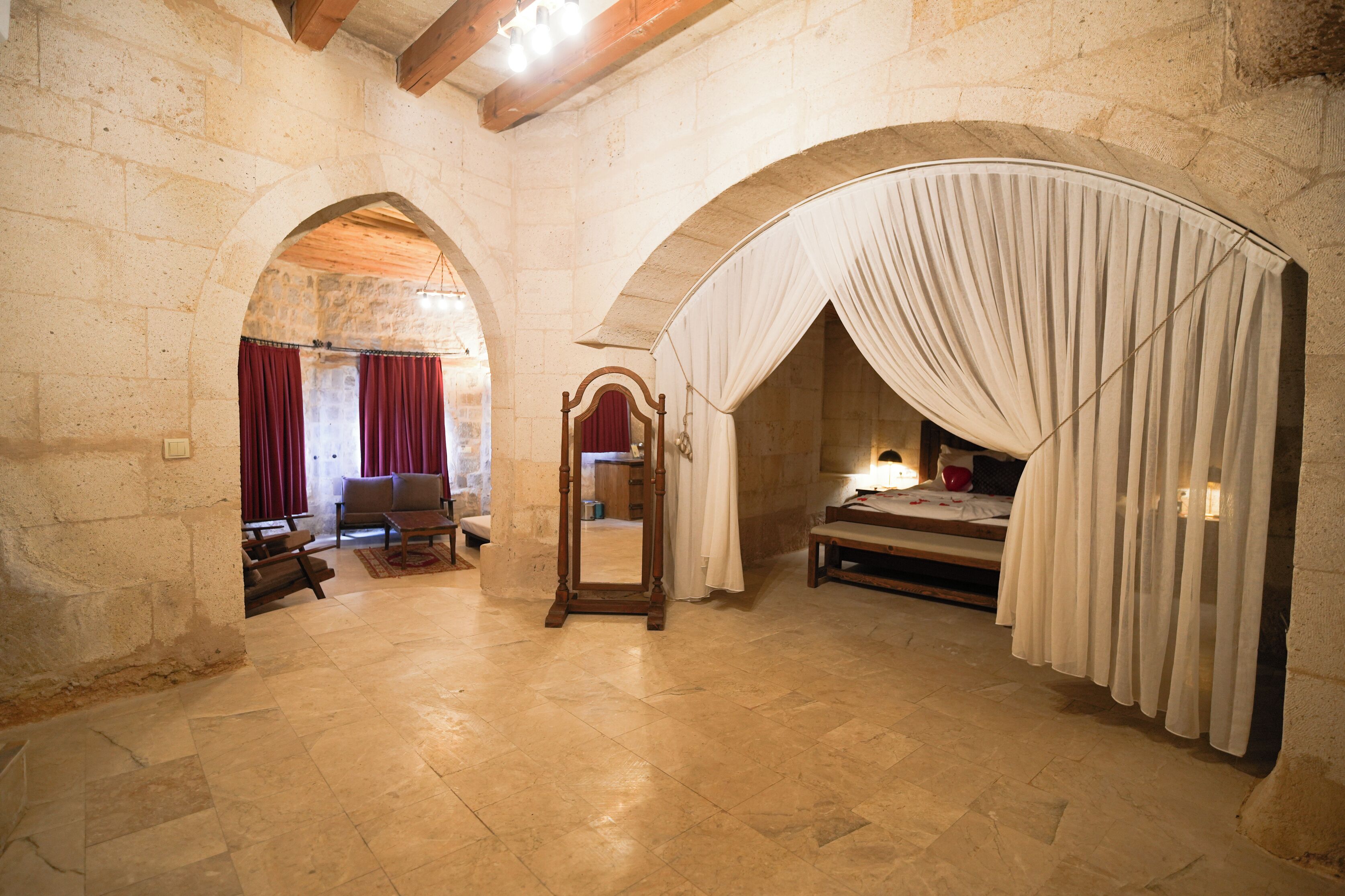 A room with stone walls, an arched doorway, a wooden bed, a nightstand, a mirror, and a lamp.