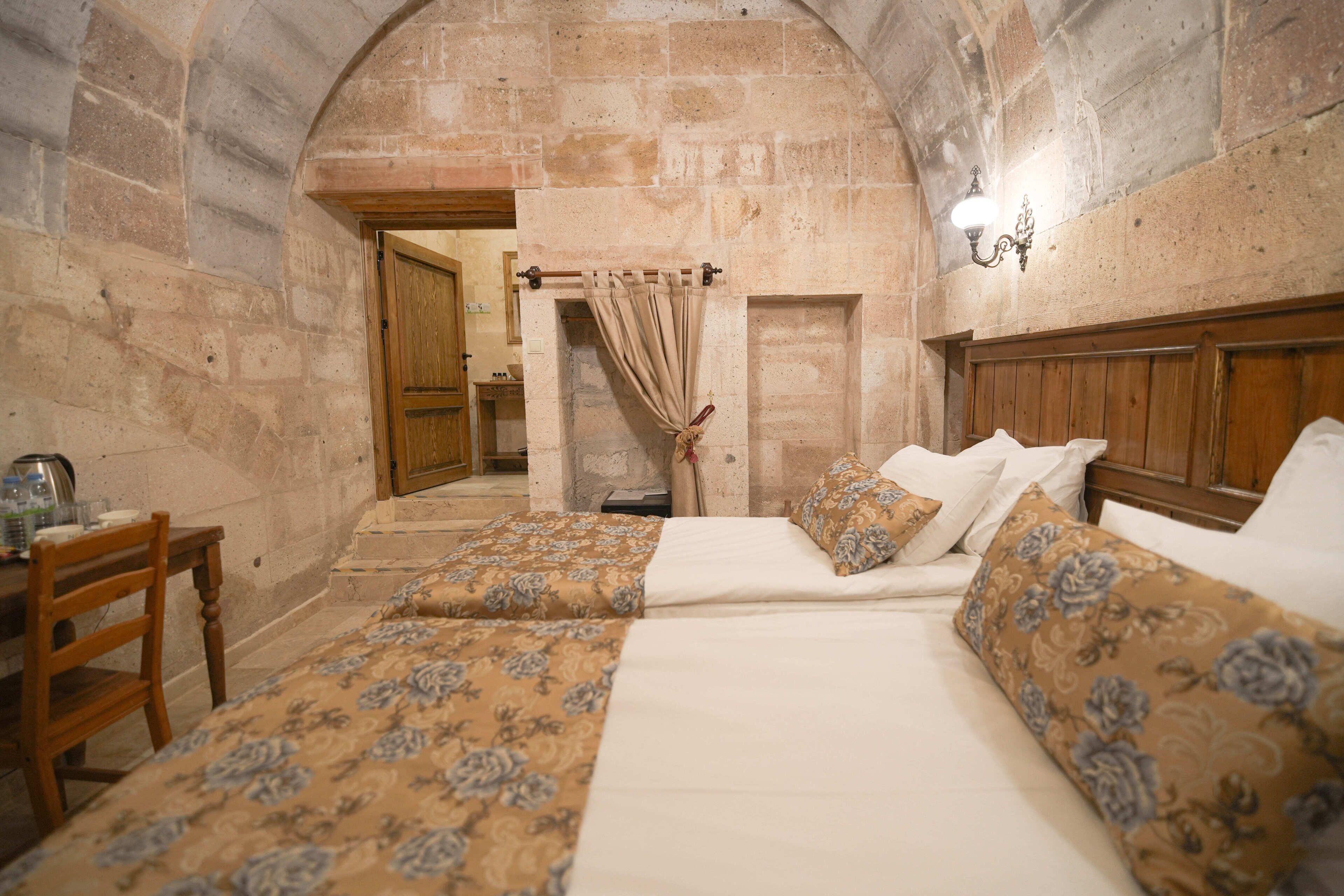 Una habitación con cama, una silla de madera, una mesa con hervidor y paredes de piedra.