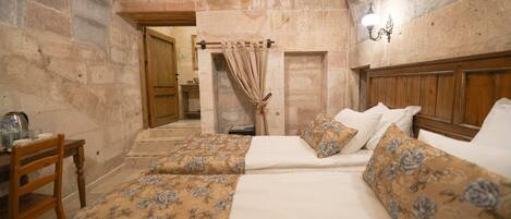 Una habitación con cama, una silla de madera, una mesa con hervidor y paredes de piedra.