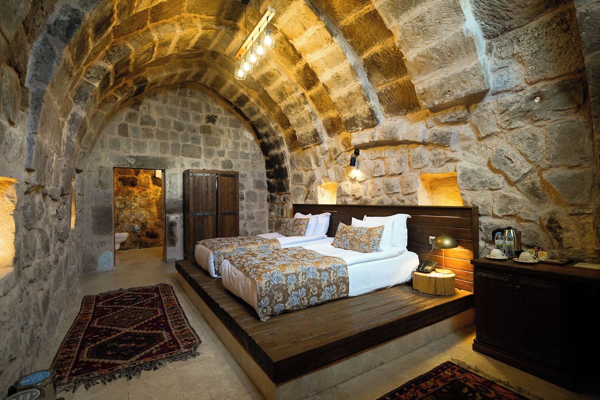 A stone-walled bedroom with a large bed, a wooden bench, and a small table with a lamp.
