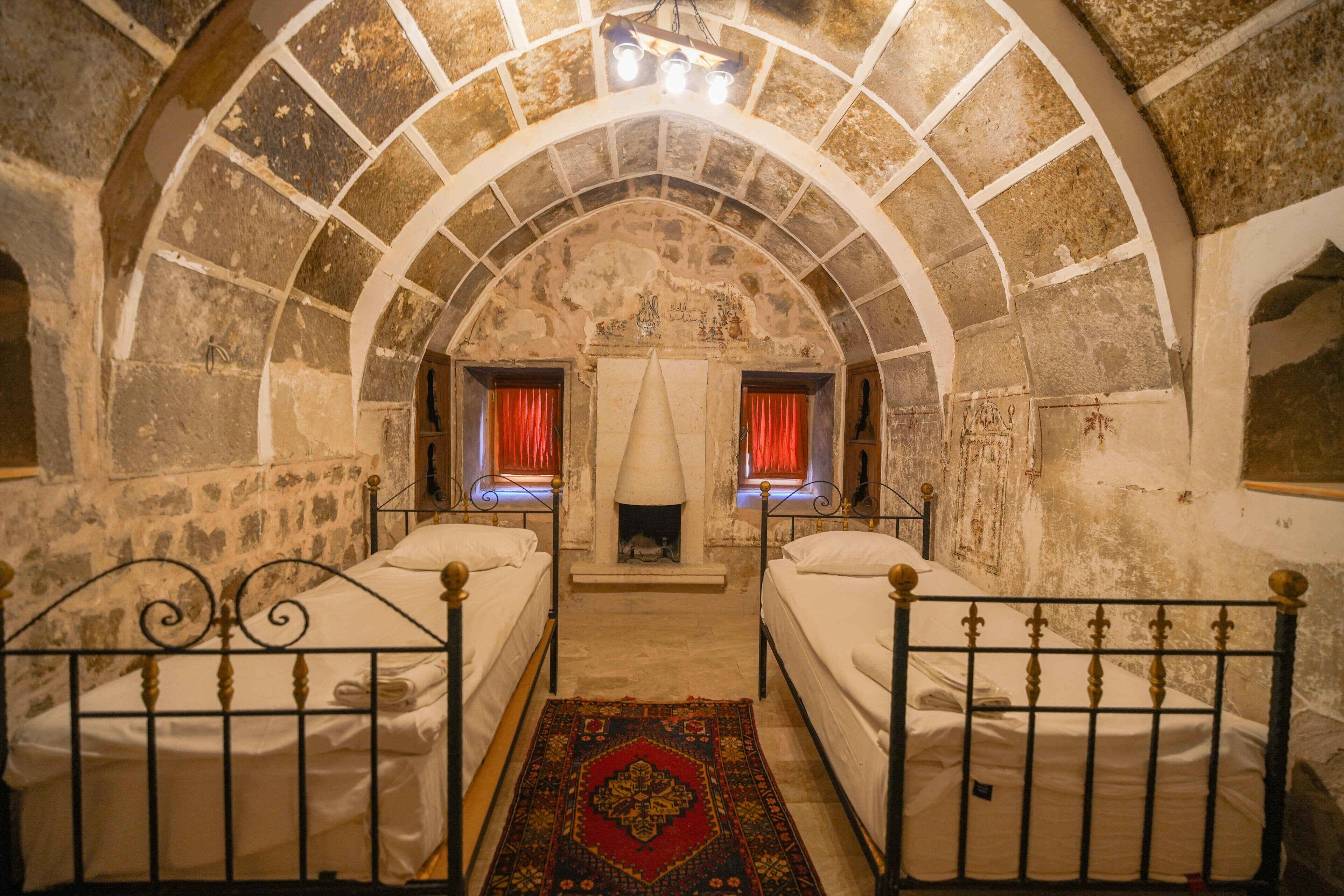A room with two beds, a stone arch ceiling, a central fireplace, and a red patterned rug.