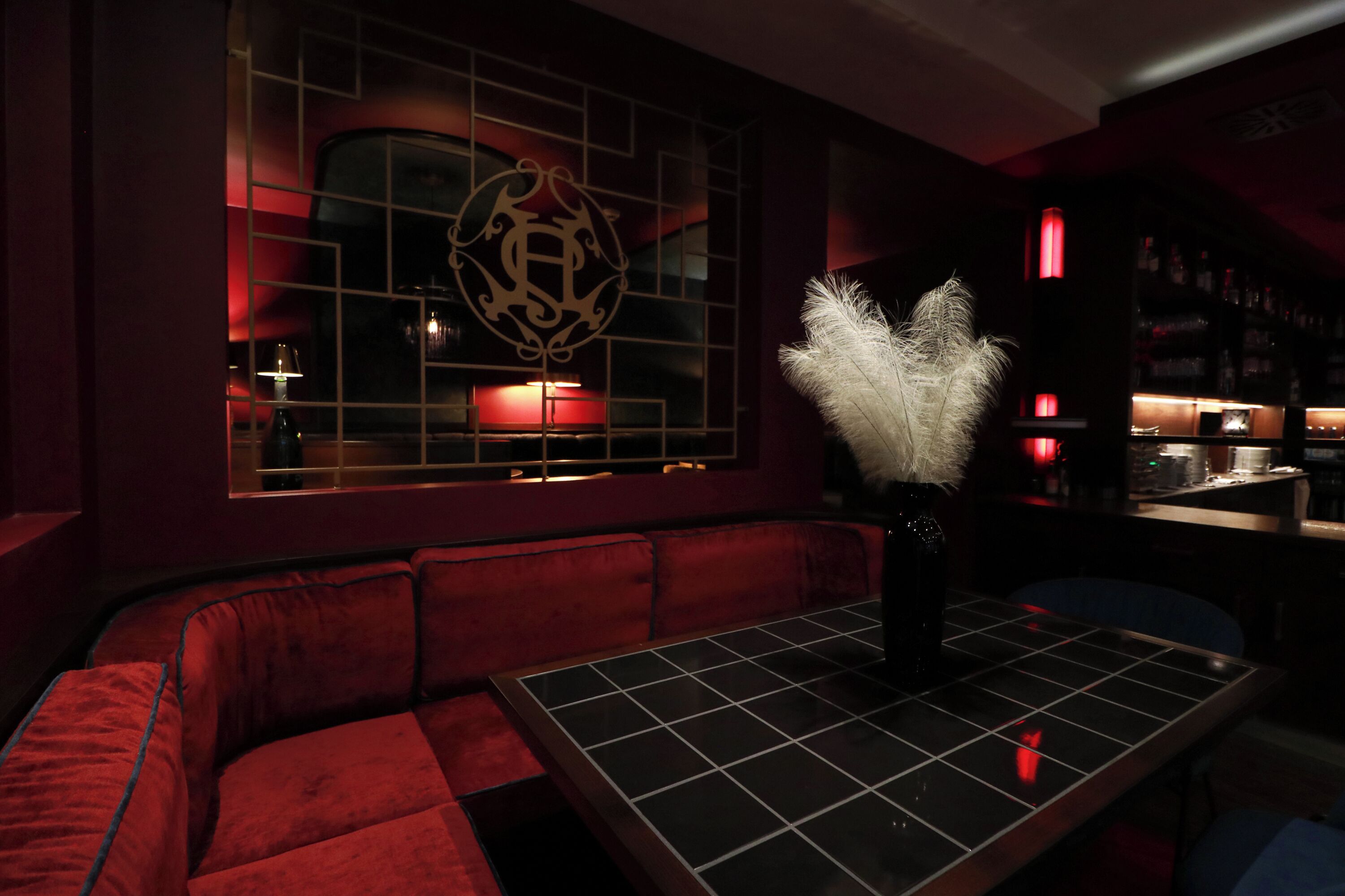 Una habitación con poca luz, un sofá tapizado en rojo, una mesa con un florero de plumas blancas y un adorno en la pared.