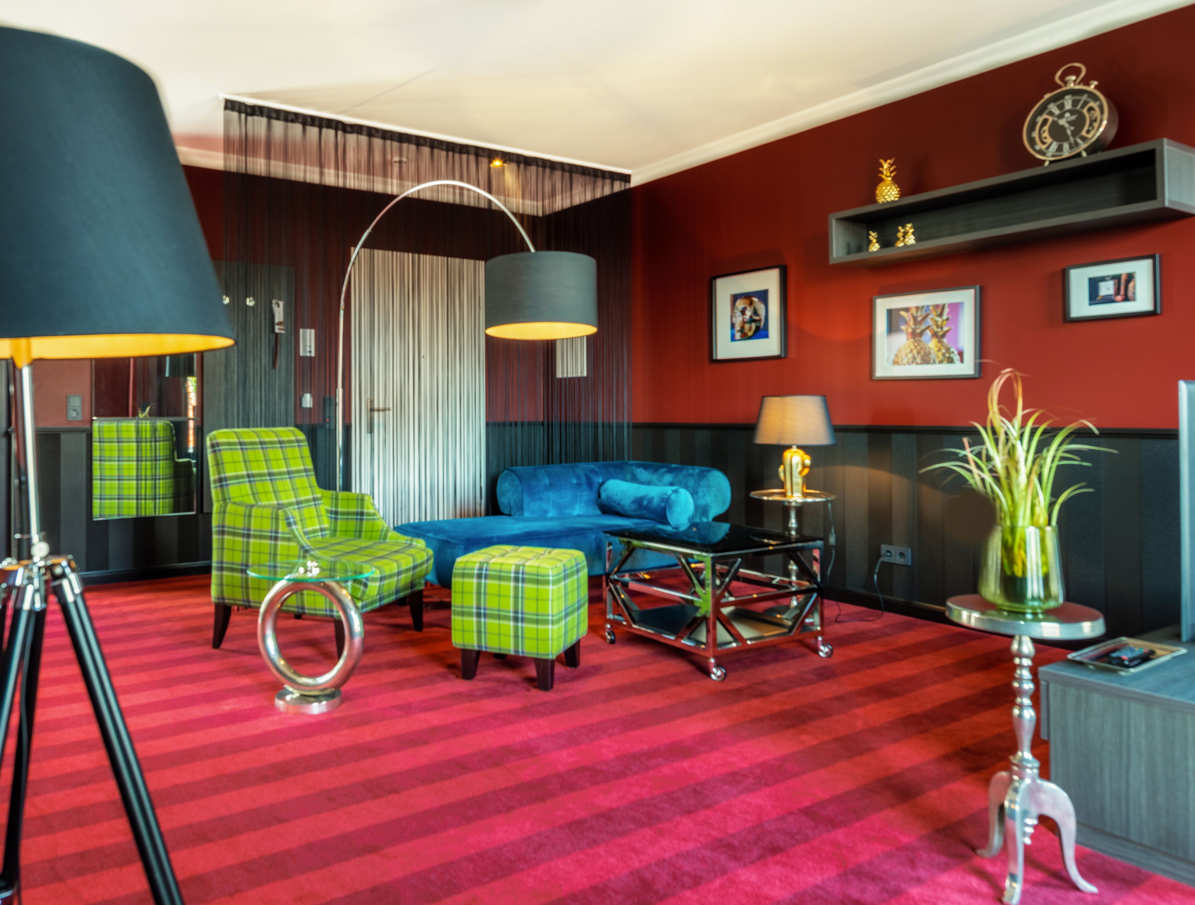 Una habitación con alfombra roja, sillones verdes a cuadros, un sofá azul, una lámpara negra y una pequeña mesa con lámpara.