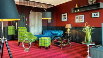Una habitación con alfombra roja, sillones verdes a cuadros, un sofá azul, una lámpara negra y una pequeña mesa con lámpara.