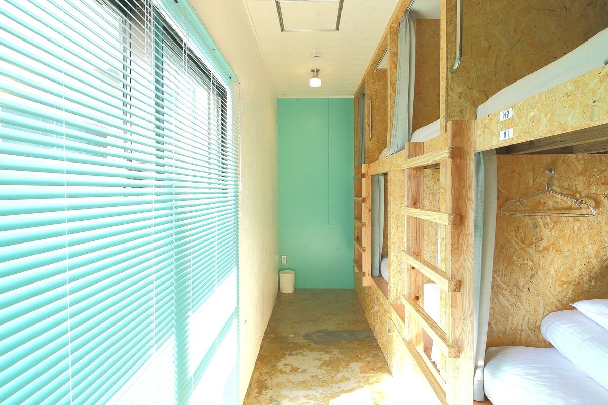 A narrow hallway with bunk beds on the right, a trash bin, and a window with blinds on the left.