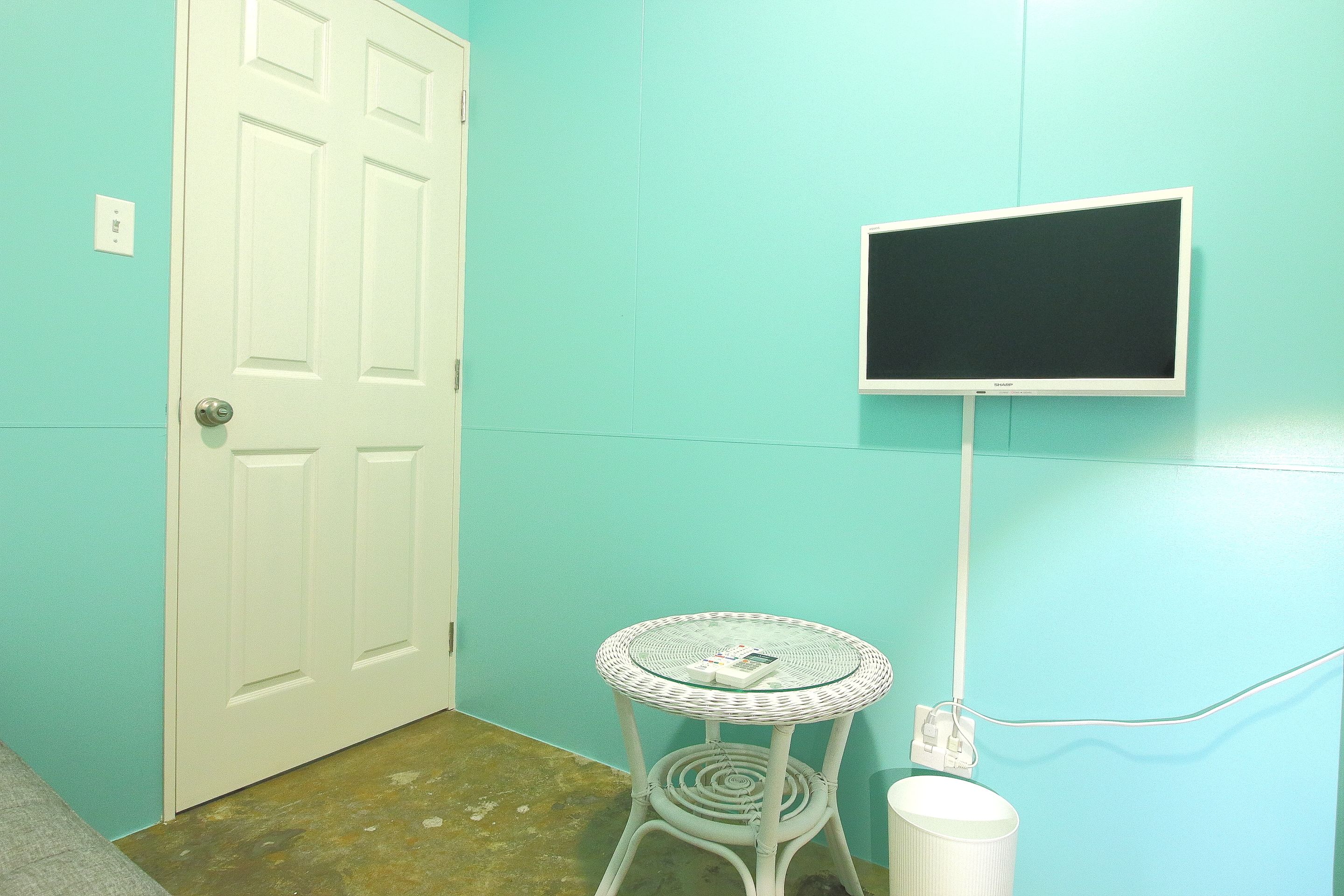 A room with teal walls, a white door, a small round table with a glass top, and a mounted flat-screen TV.