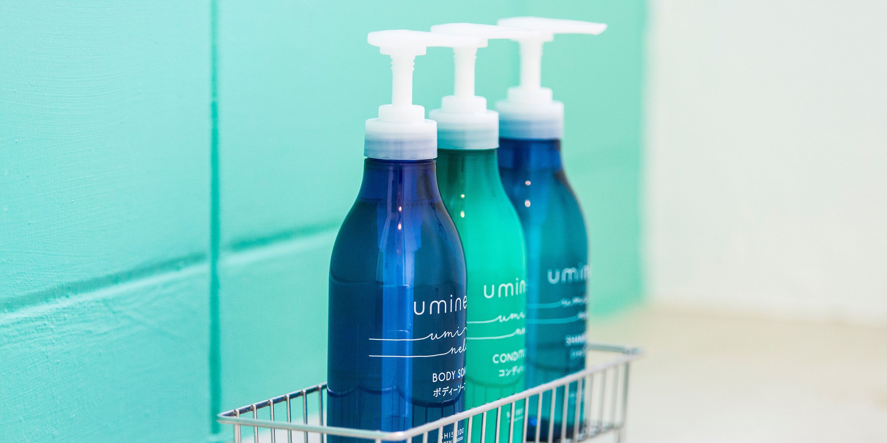 Three bottles of body soap in a metal basket against a teal wall.
