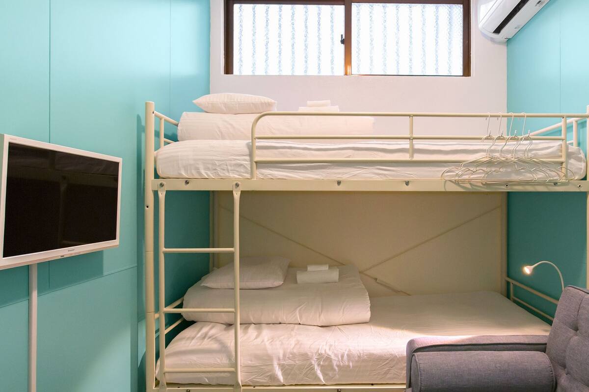 A bunk bed room with a television on the left, a window with blinds, and a chair in the foreground.