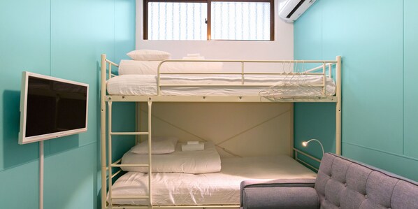 Quarto com beliches, televisão à esquerda, janela com persianas e uma cadeira em primeiro plano.