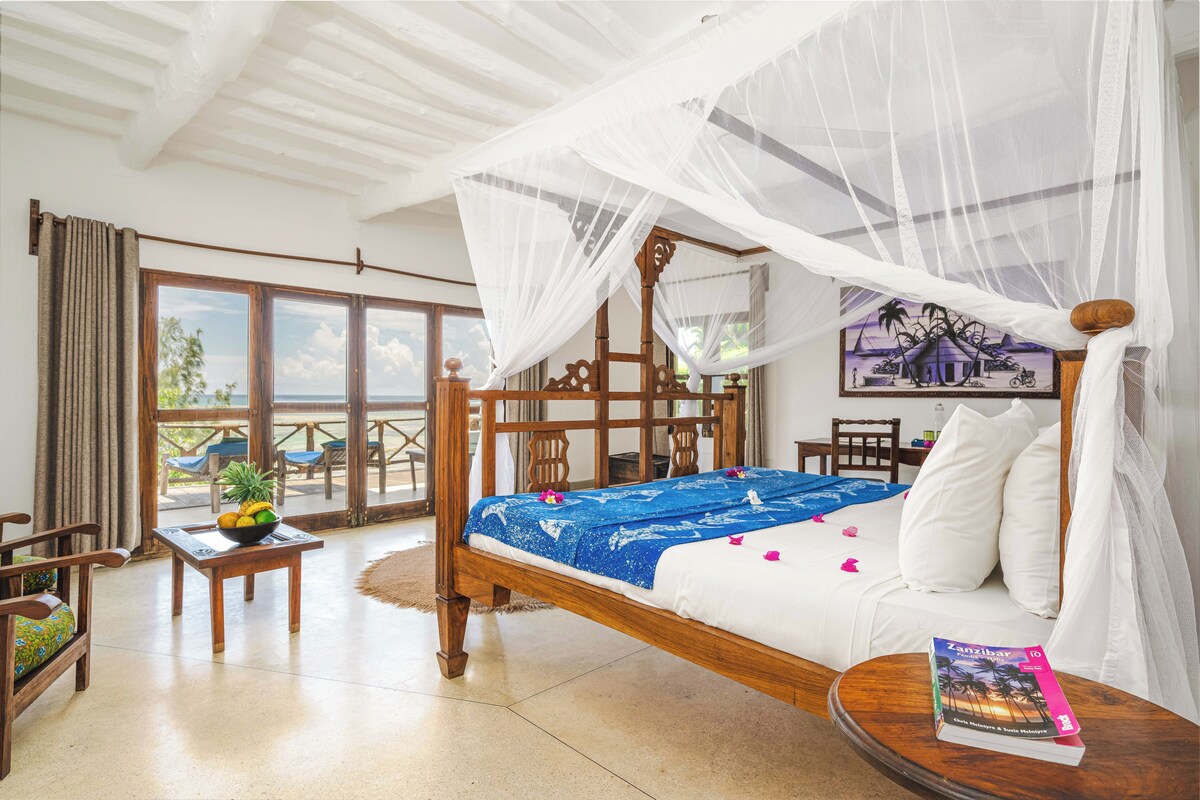 A room with a bed, a table with fruit, a chair, a balcony with a view, and a book on the bed.