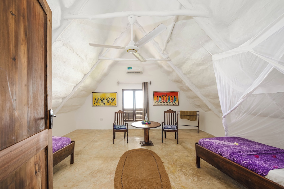 Una habitación con techo alto, una cama, una silla, una mesa y obras de arte en las paredes.