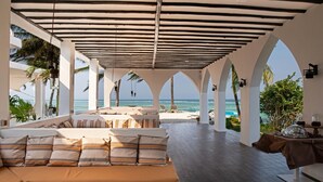 A spacious indoor area with a view of the ocean, featuring a sofa with striped cushions, a dining table with a brown tablecloth, and a wooden coffee table. - Shooting Star Boutique Hotel (Kiwengwa)