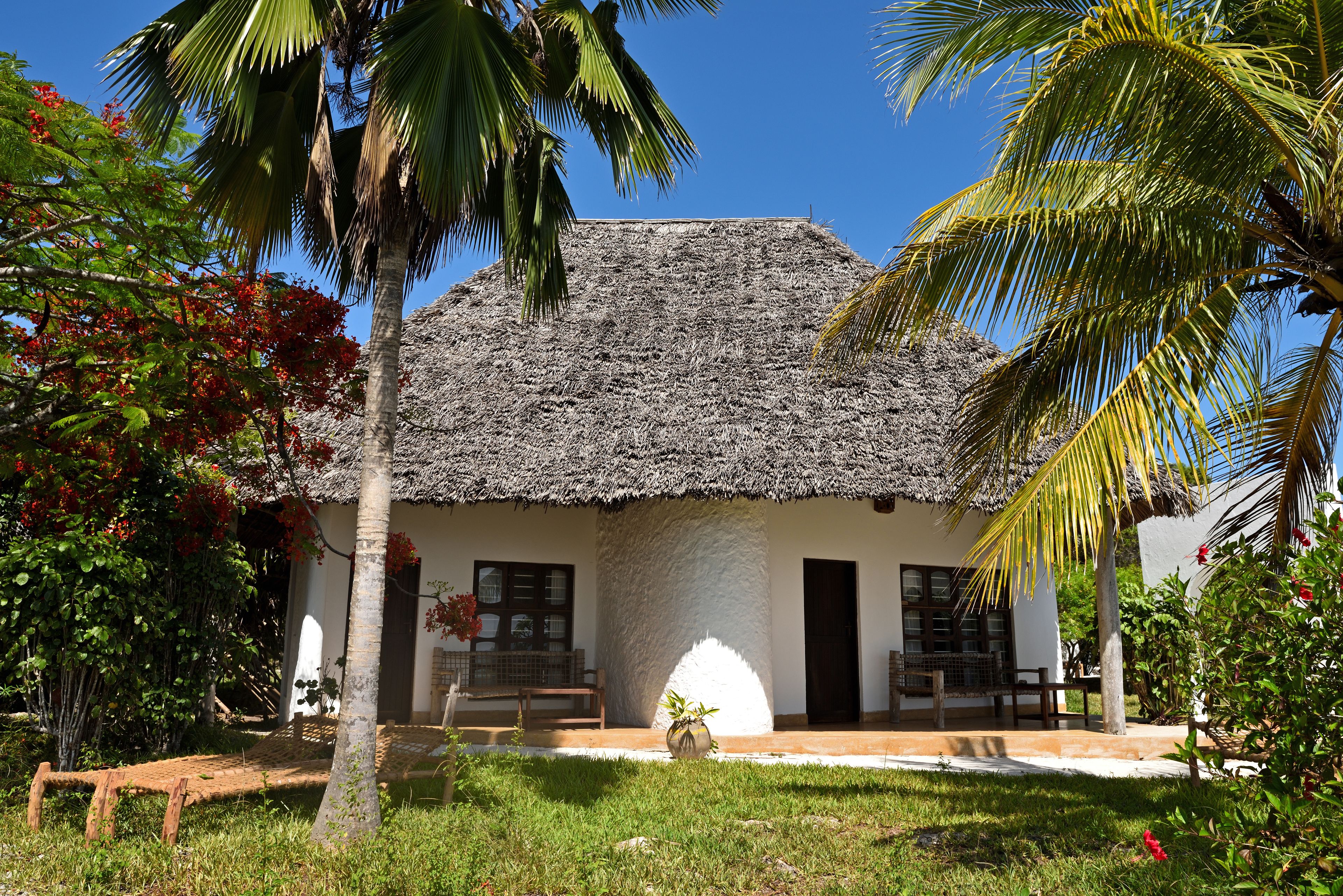 Uma casa com telhado de palha e fachada branca, um pequeno alpendre e um jardim com palmeiras e flores vermelhas.