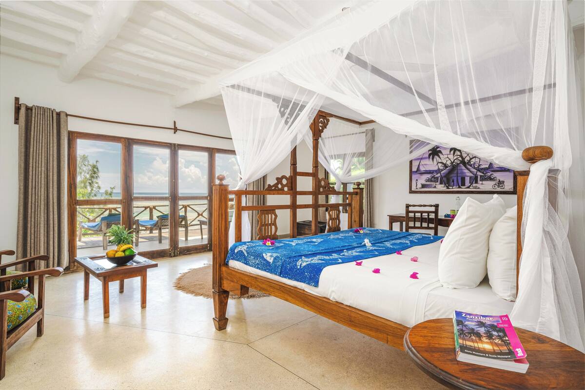 Una habitación con una cama, una mesa con fruta, una silla, un balcón con vistas y un libro sobre la cama.