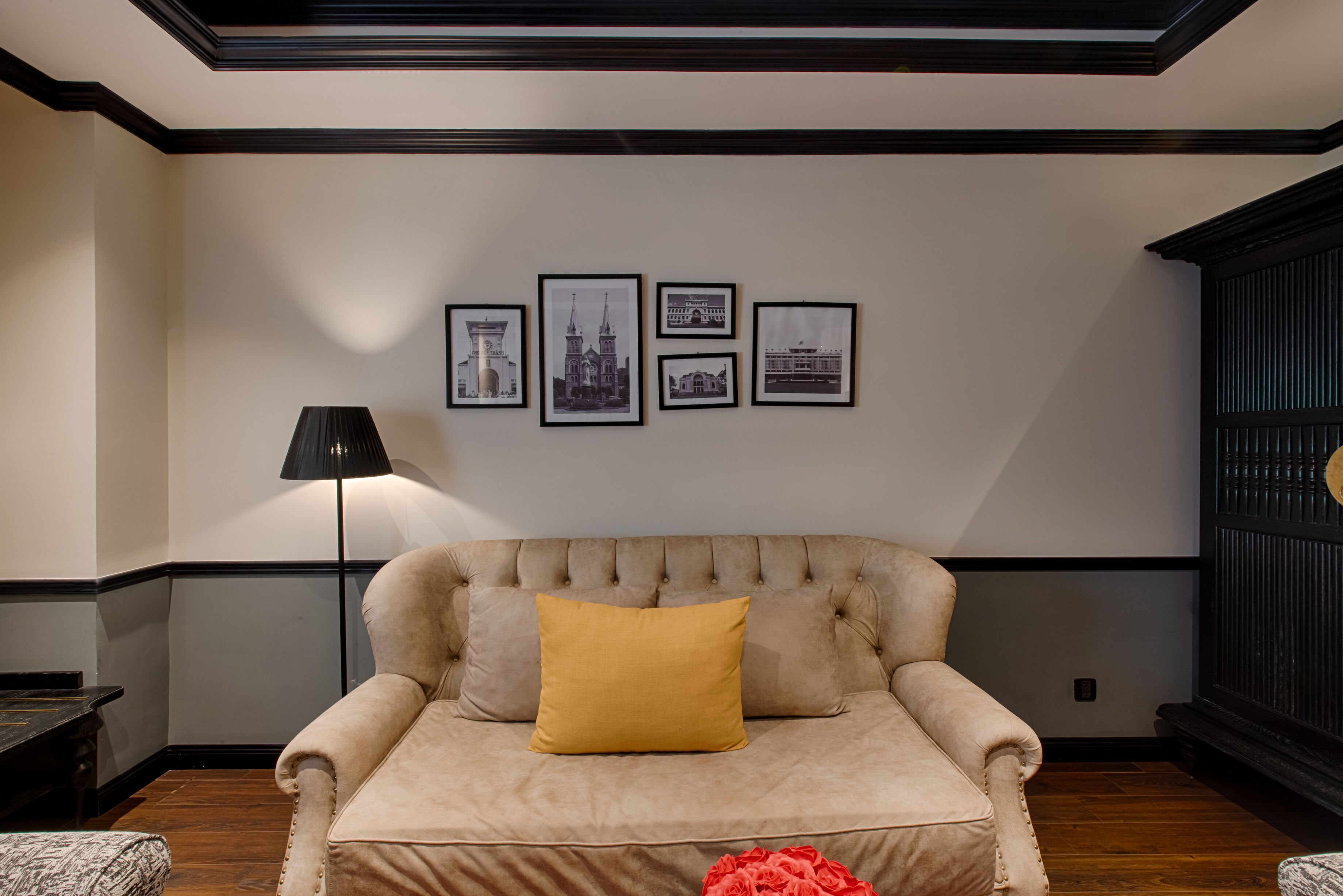 Una habitación con un sofá color beige, un cojín amarillo y una lámpara negra. Cuatro cuadros enmarcados adornan la pared.