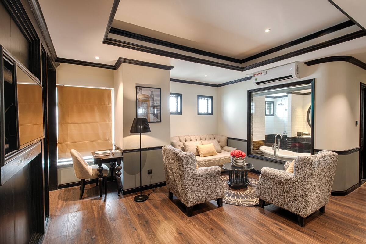 A modern living room with a sofa, two armchairs, a coffee table, and a dining table with chairs. There is a large mirror reflecting the room and a window with blinds.