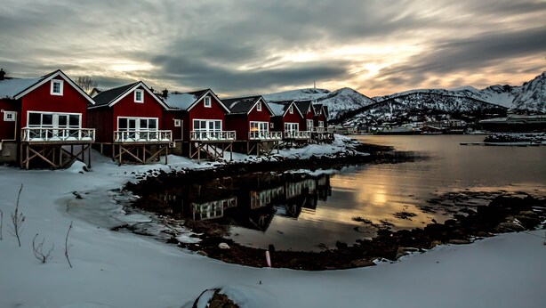 Eine Reihe roter Holzhäuser mit Balkonen, ein gefrorener See und schneebedeckte Berge im Hintergrund.
