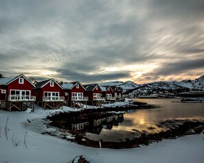 Une rangée de maisons en bois peintes en rouge, dotées de balcons, un lac gelé et, au loin, des montagnes enneigées.