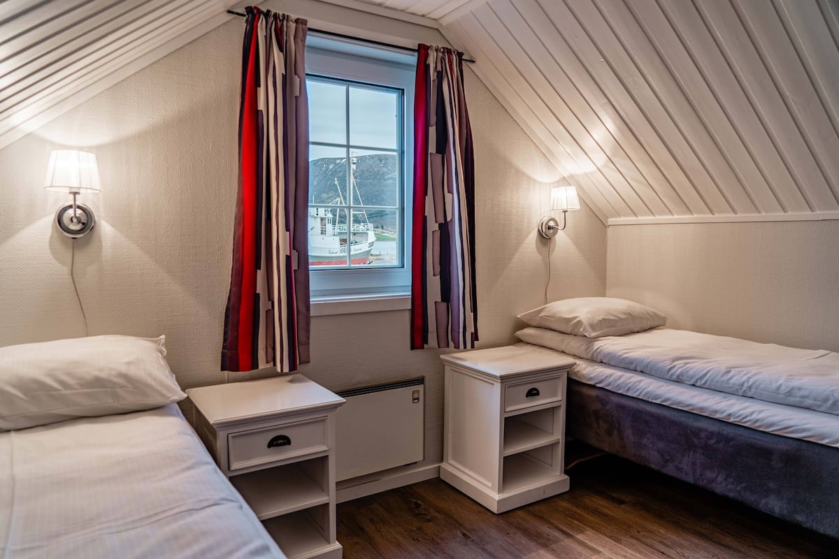 A room with a sloped ceiling, a window with curtains, two bedside tables with lamps, and a bed.