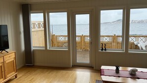 Ein Zimmer mit Holzboden, einem Fernseher auf einem Holzschrank und großen Fenstern mit Blick auf das Meer sowie einer Holzterrasse davor.
