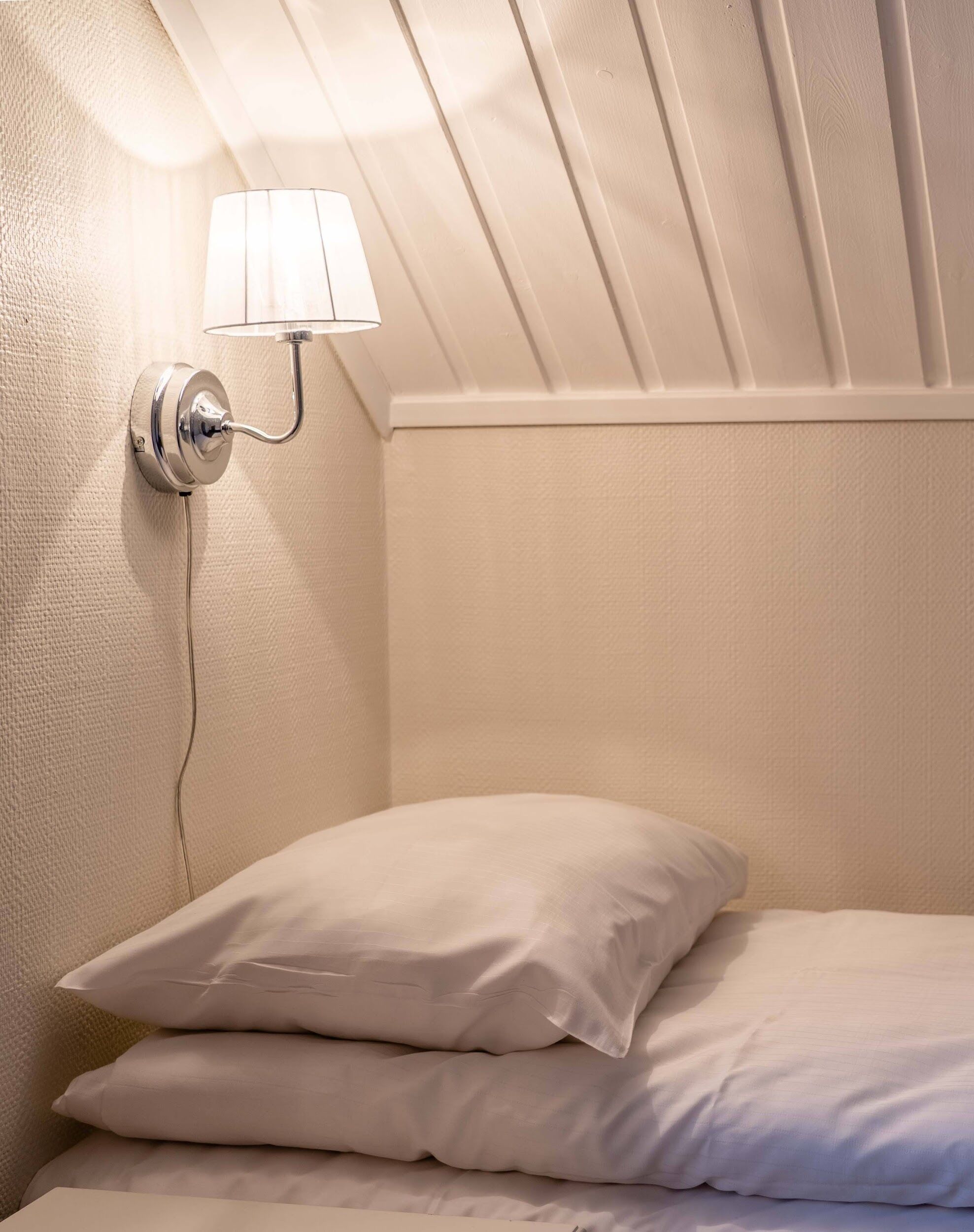 A neatly made bed with white linens and pillows, a wall-mounted light fixture, and a textured wall.