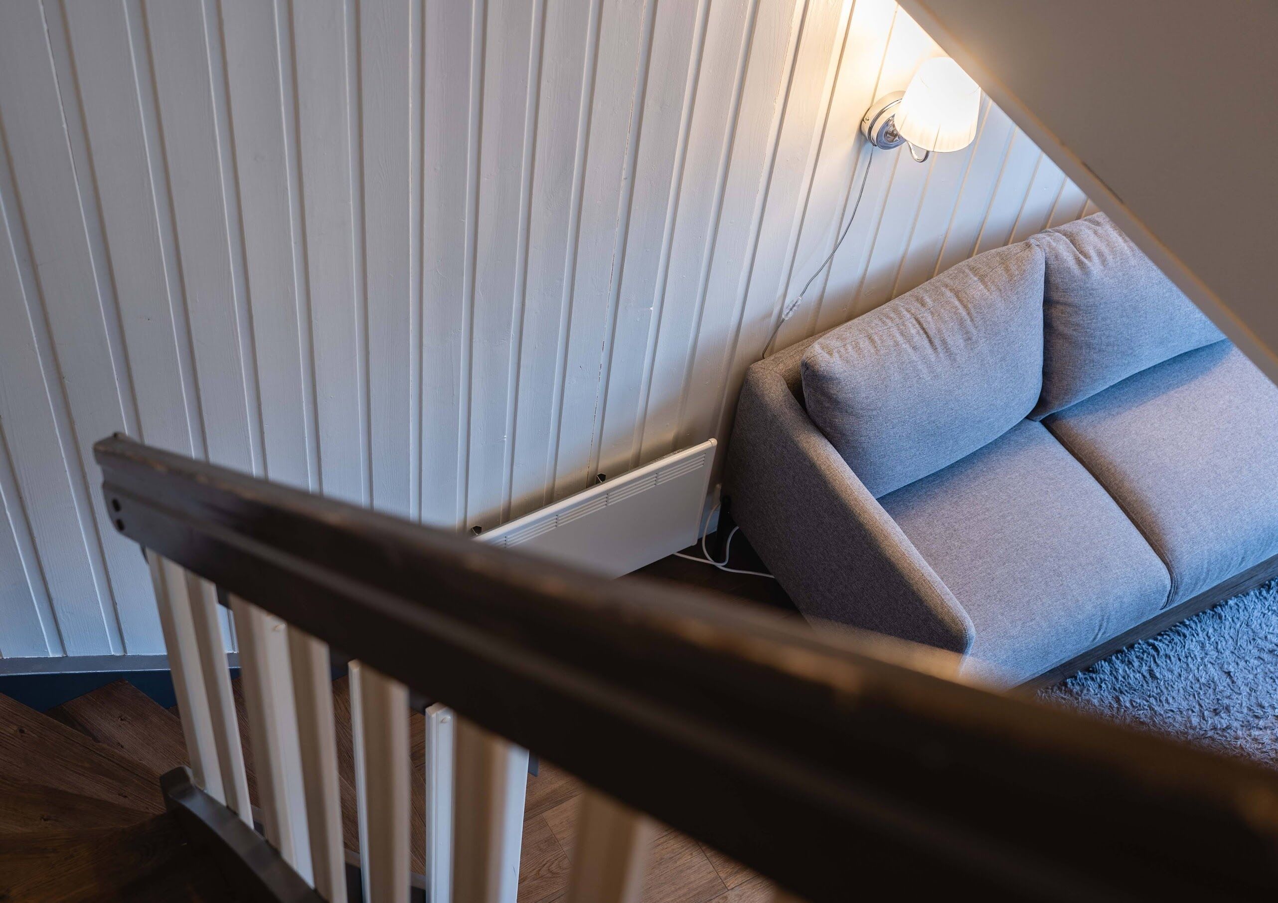 Un escalier avec une rampe en bois mène à une pièce où se trouve un canapé gris et un luminaire fixé au mur.
