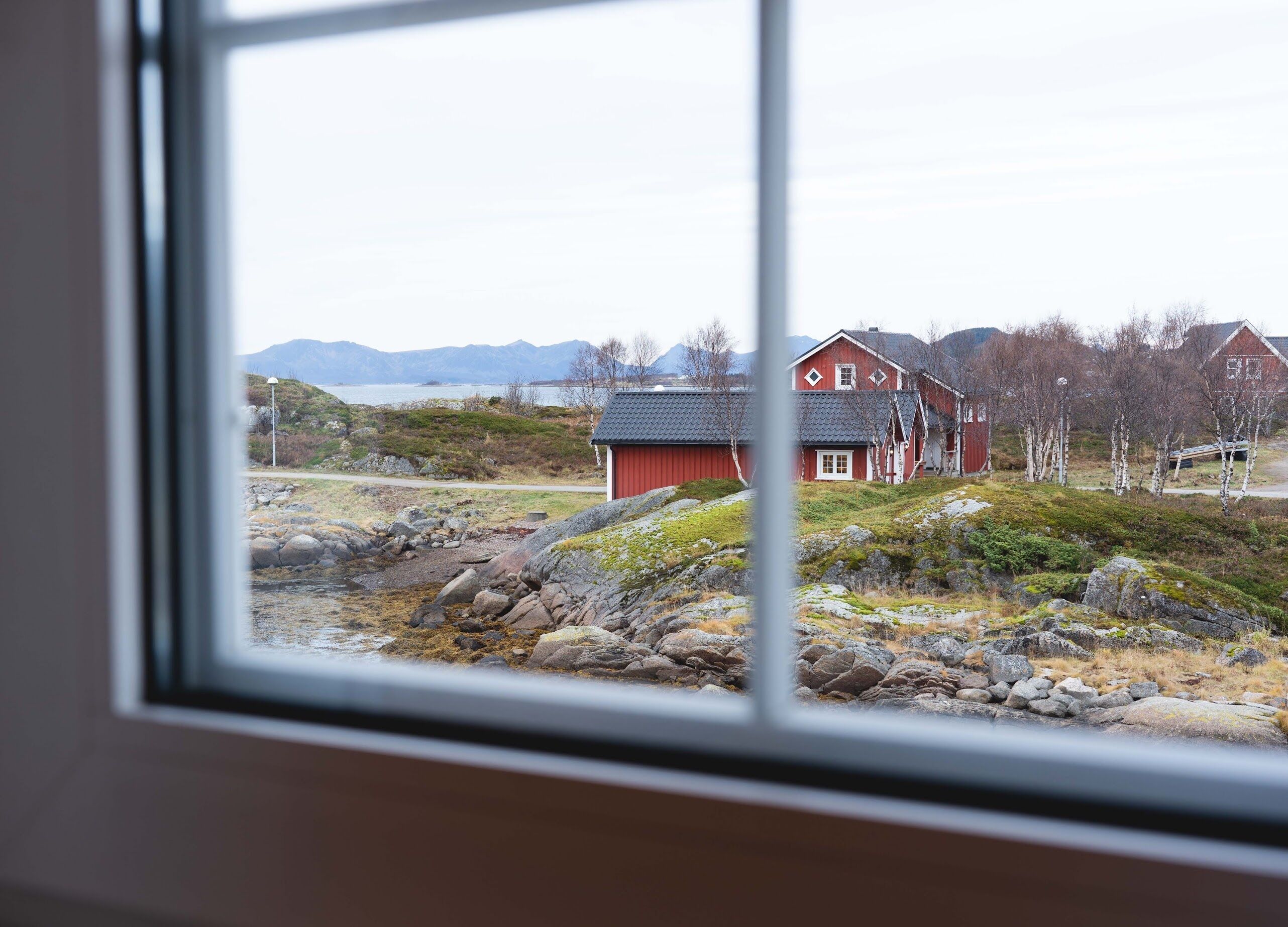 A red house with a white door and a small porch, surrounded by a rocky landscape and mountains in the background.