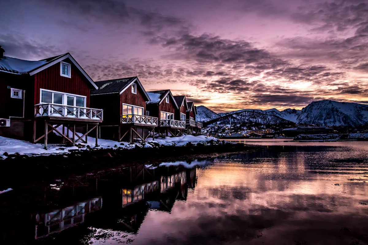 A row of red wooden houses with balconies, a calm lake, and snow-covered mountains in the background.